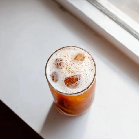 A tall glass of Iced Chai Tea with Cold Foam layered over ice cubes, crowned with creamy foam and a light dusting of cinnamon.